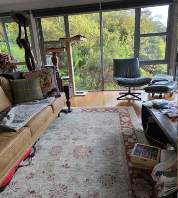 Ein gemütliches Wohnzimmer mit einem hellbraunen Sofa, Zierkissen, einem gemusterten Teppich, einem Kratzbaum, großen Fenstern, Grünpflanzen draußen, einem modernen Sessel und hier und da verstreuten Bastelmaterialien. Warme Atmosphäre.