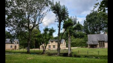 This image shows a peaceful countryside scene with three quaint houses, tall leafy trees, lush green grass, a small stream, wooden fence, and a partly cloudy sky.