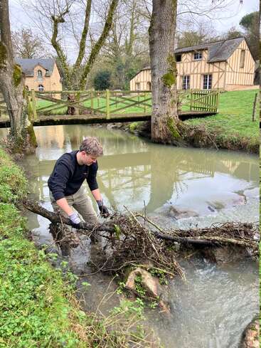 Jemand mit Handschuhen räumt Äste und Müll aus einem kleinen Bach, mit rustikalen Häusern, einer Holzbrücke, Bäumen und grasbewachsenen Ufern im ruhigen Hintergrund.