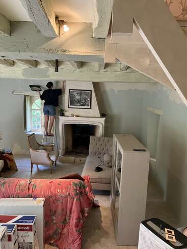 Una persona está de pie sobre una silla pintando las paredes junto a una chimenea en una habitación acogedora y desordenada, con vigas a la vista, muebles y una manta de flores. Se están realizando obras de reforma.
