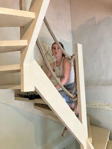 Una mujer vestida con una camiseta sin mangas blanca y vaqueros está sentada en unas escaleras de color beige con barandillas de cuerda gruesa, en una habitación con paredes texturizadas y parcialmente pintadas.