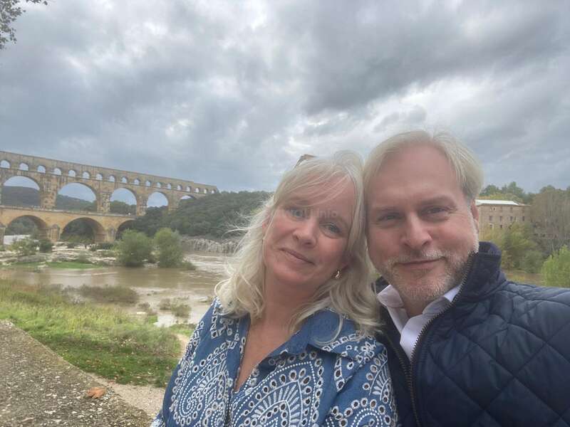 A smiling couple takes a selfie outdoors. Behind them stands an ancient stone aqueduct, a river, greenery, and dramatic cloudy skies, capturing a peaceful, scenic moment.