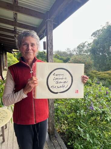 A smiling woman stands on a porch surrounded by greenery, holding a sign reading, “happy farmers change the world,” with a black circular brushstroke around the text.