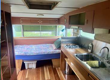 This cozy camper interior features a bed with colorful bedding, wooden flooring, a compact kitchen with a stove and sink, cabinets, and large windows.
