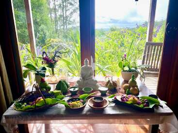 Un sereno altar interior cuenta con una estatua de Buda, flores vibrantes, ofrendas de fruta y cuencos, vegetación y luz natural que entra por grandes ventanas con vistas a un exuberante jardín.