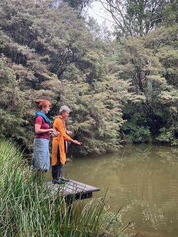 Dos mujeres están de pie sobre una pequeña plataforma de madera junto a un estanque tranquilo, rodeadas de frondosos árboles verdes y juncos, aparentemente compartiendo un momento de paz y contemplación.