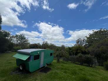 Una caravana vintage verde descansa sobre una exuberante pradera rodeada de árboles bajo un cielo azul brillante con nubes dispersas, creando una atmósfera tranquila y pintoresca de campo.