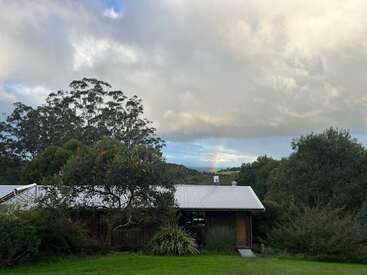 Una pequeña casa con techo de chapa se encuentra entre frondosos árboles y vegetación, bajo un cielo nublado, con un tenue arco iris visible en las colinas lejanas.