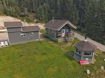 This image shows three small grey buildings, including a cabin, on a grassy lawn surrounded by forest. A few white chairs and paths can be seen.