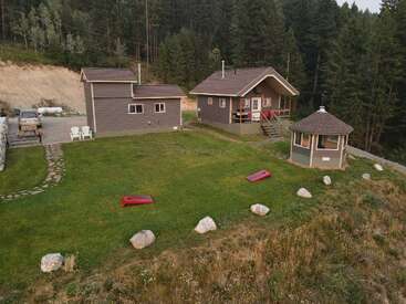 A cozy cabin sits in a lush green yard with a small outbuilding, gazebo, lawn games, stone path, surrounded by forest and tranquility. Peaceful setting.