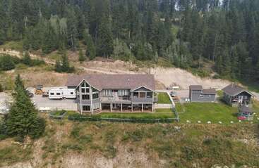 This image shows a large house with a sloped roof in a clearing, surrounded by dense forest, smaller buildings, a camper, and a grassy yard.