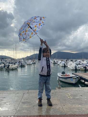 Ein lächelndes Kind steht in der Nähe eines Jachthafens und hält einen bunten Regenschirm in die Höhe. Im Hafen dahinter liegen Boote unter einem dramatisch bewölkten Himmel vor Anker.