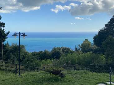 Eine malerische Aussicht auf das Meer, mit üppig grünen Bäumen und Gras im Vordergrund, einer alten Straßenlaterne, einem Holzzaun und einer Schubkarre.