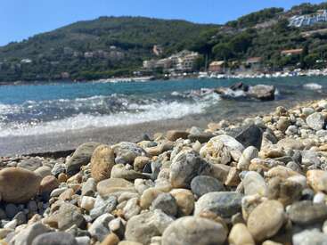 Ein mit Kieselsteinen übersäter Strand im Vordergrund, sanfte Wellen nähern sich. Das blaue Meer, die Hügel in der Ferne und die Gebäude an der Küste unter einem klaren Himmel schaffen eine ruhige mediterrane Szene.