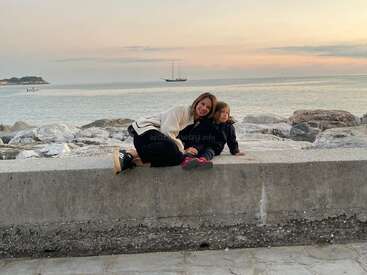 Eine Frau und ein kleines Kind sitzen bei Sonnenuntergang auf einer Steinmauer am Meer, lächeln und entspannen sich, während in der Ferne ein Segelboot zu sehen ist.