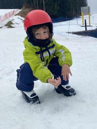 Ein kleines Kind in grüner Jacke, blauer Hose und rotem Helm hockt auf dem verschneiten Boden und hält den Schnee fest, bereit für ein Winterabenteuer im Freien.