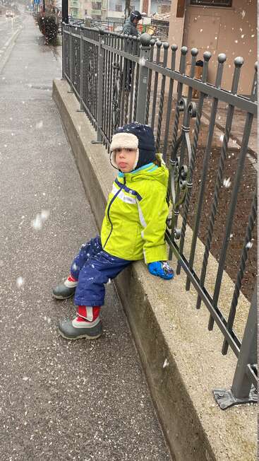 Ein kleines Kind in Winterkleidung sitzt auf einer niedrigen Mauer an einem Metallzaun und streckt seine Zunge heraus, während um es herum sanft Schnee fällt.