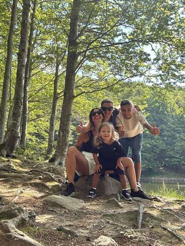 Eine glückliche vierköpfige Familie posiert gemeinsam in einem sonnenbeschienenen Wald und lächelt in die Kamera. Bäume, Wurzeln und grünes Laub umgeben sie und schaffen eine friedliche Atmosphäre.