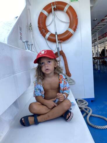 Ein kleines Kind sitzt im Schneidersitz auf einem Boot und trägt eine rote Mütze und ein geblümtes Hemd. Hinter ihnen sind eine orangefarbene Rettungsweste und ein aufgerolltes Seil zu sehen.