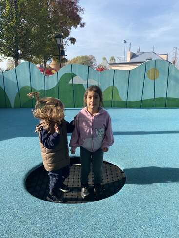 Zwei Kinder stehen auf einem Trampolin auf einem Spielplatz. Die Haare des einen Kindes wehen im Wind, während das andere direkt in die Kamera schaut und leicht lächelt.