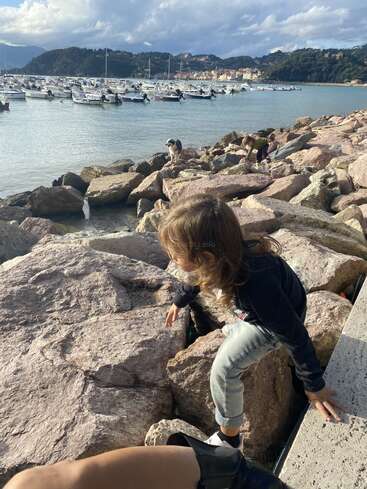 Ein kleines Kind klettert auf große Felsen am Meer, Boote liegen im Wasser vor Anker, eine entfernte Stadt, Berge und ein Hund sind ebenfalls zu sehen.