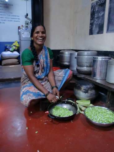 Das Bild zeigt eine Frau in einer Küche, die auf dem Boden sitzt und Gemüse in einer Pfanne zerkleinert. Sie trägt einen Sari und hat eine Uhr an ihrem linken Handgelenk.