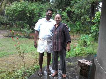 Das Bild zeigt zwei Männer, die auf einer steinernen Terrasse in einem üppigen Garten stehen, umgeben von Bäumen und Gras, wobei ein Mann ein weißes und der andere ein dunkles Hemd trägt.