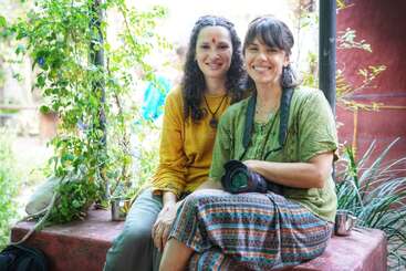 Two women sit together outdoors, smiling warmly. One wears bright patterned clothes and holds a camera, while the other wears yellow, both surrounded by lush greenery.