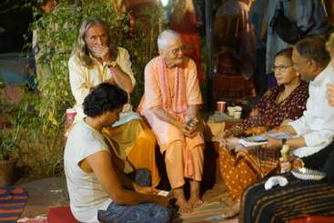 A group of people sit together outdoors at night, engaged in conversation. Some hold papers, notebooks, or snacks, creating a relaxed, communal, and spiritual atmosphere.