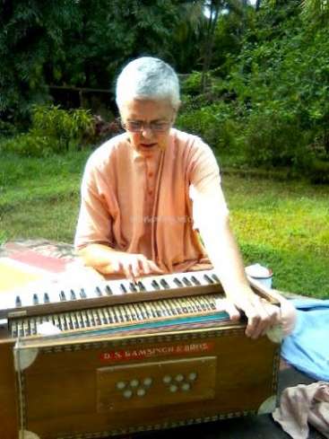 Eine ältere Person mit kurzen weißen Haaren, die eine pfirsichfarbene Kurta trägt, spielt im Freien, umgeben von grünen Bäumen und Sonnenlicht, ein Harmonium und wirkt konzentriert und gelassen.