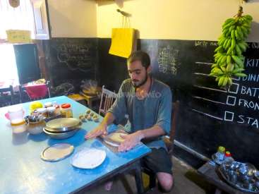 Das Bild zeigt einen Mann, der auf einem blauen Tisch in einer Küche Teig knetet, umgeben von verschiedenen Zutaten und Kochutensilien.