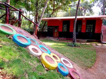 Das Bild zeigt ein leuchtend rotes Gebäude mit einer abfallenden Rasenfläche, auf der ein bunter Reifenweg zum Eingang führt. Das Gebäude ist von üppigen Bäumen umgeben.