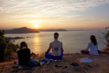Das Bild zeigt drei Personen, die auf einem Hügel sitzen und ein Gewässer überblicken, während in der Ferne die Sonne untergeht.