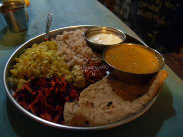 Das Bild zeigt einen Metallteller mit verschiedenen Lebensmitteln, darunter Reis, Gemüse und Fladenbrot, die auf einem Tisch angeordnet sind.