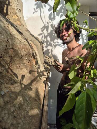 Un homme torse nu sourit en travaillant avec de l'argile, en construisant ou en réparant un mur en terre. La lumière du soleil et les feuilles vertes le recouvrent partiellement, créant des ombres naturelles sur le mur.