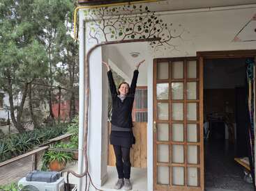 Une femme se tient dans l'embrasure d'une porte, les bras levés, souriante. Une fresque d'arbre est peinte au-dessus d'elle, la faisant ressembler à une partie de l'arbre avec ses branches.