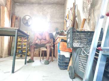 Um homem sem camisa senta-se em uma cadeira de madeira em uma oficina desordenada e iluminada pelo sol, usando crocodilos verdes, cercado por ferramentas, caixas de armazenamento e materiais de madeira inacabados.