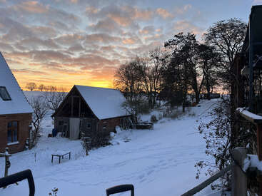 A snowy countryside scene with houses, trees, and a soft orange sunset sky. Fresh snow covers the ground and rooftops, creating a peaceful winter atmosphere.