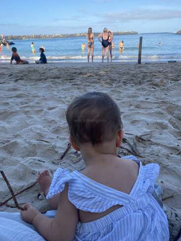 A baby in a blue striped outfit sits on the sandy beach, watching people swimming and playing in the ocean under a bright, clear sky.