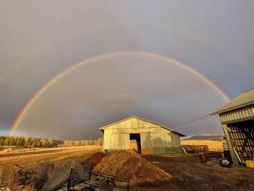 A vibrant double rainbow arcs across a cloudy sky over a rustic farm. Metal barns, hay, and an open field add charm to the tranquil rural scene.