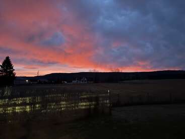 A dramatic sunset sky glows orange and pink above a rural landscape. Dark hills, scattered houses, a fence, and field grass are visible in the foreground.