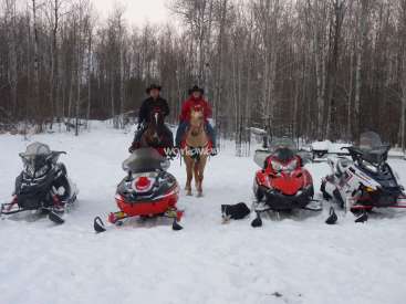 L'image représente deux personnes à cheval dans un paysage enneigé, entourées de motoneiges et d'un chien. La scène se déroule sur fond d'arbres.