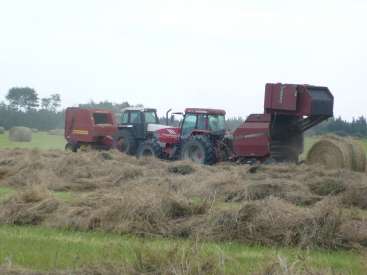 L'image représente un tracteur et une presse à balles dans un champ, entouré de foin, avec des arbres visibles à l'arrière-plan par une journée nuageuse.