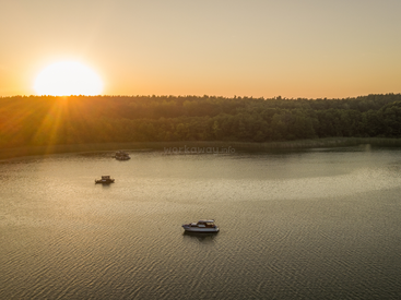 Um pôr do sol tranquilo sobre um lago calmo, três barcos flutuando suavemente na água, uma floresta verdejante no horizonte, a luz dourada do sol refletindo lindamente. Uma noite pacífica.