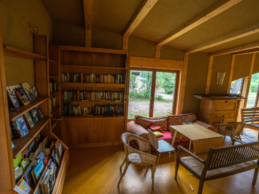 Esse quarto aconchegante tem estantes de madeira cheias de livros, um sofá colorido, cadeiras de vime, uma mesa de madeira e janelas grandes com vista para um jardim verde e tranquilo.