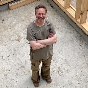 The image depicts a man standing in a construction site, with wooden framing surrounding him, wearing a gray t-shirt and khaki pants, arms crossed, and smiling at the camera.