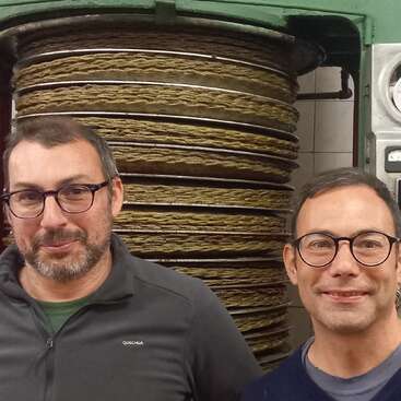 Dos hombres con gafas sonríen delante de una gran máquina industrial con bandejas circulares apiladas llenas de un material fibroso y texturizado en una sala alicatada.