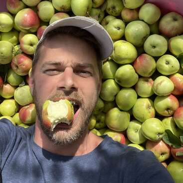 Un hombre con barba y gorra blanca está tumbado sobre un montón de manzanas. Se está haciendo un selfi mientras muerde alegremente una manzana fresca.