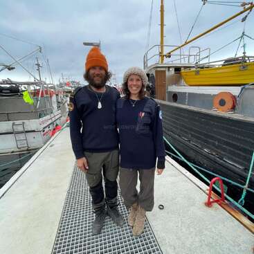 The image depicts a man and woman standing on a dock, surrounded by boats, with the man wearing an orange beanie and the woman sporting a beige hat.