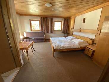 This cozy room features a wooden bed, side tables, a small table with chairs, a sofa, two windows with curtains, and warm wooden accents throughout.